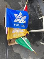 Lot of 5 Canadian Centennial flags in various colors piled together, showing blue, yellow, and green flags with white maple leaf Centennial design and the years 1867 | 1967, each attached to a stick with a gold spear tip.