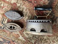 Antique charcoal iron with black wooden handle placed on a patterned fabric background along with two sad iron inserts.