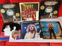 Collection of six vintage rock and disco vinyl albums displayed on a red couch, featuring covers of Saturday Night Fever, Free, Frampton Comes Alive!, Elton John, and The Rolling Stones.
