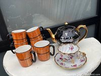 Set of one black teapot with gold trim, one white footed tea cup with flower design and matching saucer, and four brown mugs with black handles.