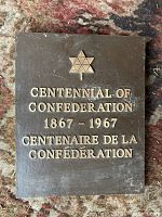 Front view of vintage metal plaque showing raised bilingual text and emblem commemorating Canadian Centennial 1867-1967 on a brown background.