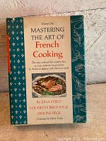 Front cover of the book showing title: Volume One Mastering the Art of French Cooking by Julia Child, Louisette Bertholle, Simone Beck. Includes color drawing of a prepared meal. Good condition dust jacket with decorative border.