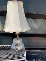 Vintage floor lamp showing entire lamp with iridescent glass base and white fabric lampshade with scalloped edge and braided trim.
