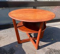 Full view of round teak coffee table showing top, lower shelf, and cross base