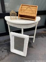 Full view of the white wicker half-moon side table with the wicker framed mirror leaning against it and the wicker stationery holder and metal wire house decor placed on top.