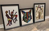 Three framed mixed media art pieces by Uti Ndueze showing different subjects: human figures, colorful mask, robed figures.