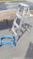 Full view of aluminum 3-step ladder showing all steps and top platform with paint marks and wear, placed on pavement next to wooden table.