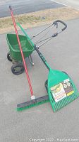 Photo showing fertilizer spreader with green hopper and partnered rake broom with red handle and green bristles