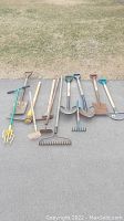 Various used garden hand tools arranged on pavement, including shovels, rakes, hoes, and cultivators with wooden and metal handles.