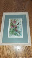 Image showing full framed artwork with hummingbirds and fuchsia flowers, cream wood frame and blue matte border over wooden surface.