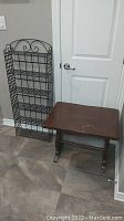 Both the black metal wire shelf and brown wooden side table shown beside a white door on tiled flooring.