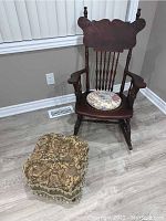 Wooden rocking chair with rounded armrests, spindle back and carved crest top, featuring a small round floral cushion at the seat, shown beside the upholstered ottoman.