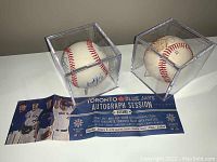 Both autographed baseballs displayed in their acrylic cases with the Toronto Blue Jays autograph session flyer and ticket beneath them.