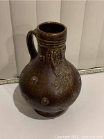 Full view of the dark brown ceramic vase with a handle, bulbous base, and cylindrical neck, featuring raised circular decorations and a bearded face motif on the neck.