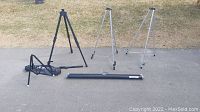 Overview of three tripods and black cylindrical projector screen case on pavement.
