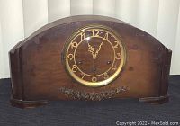 Front view of wooden mantel clock with clock face showing numbers and hands, decorative carving below face.