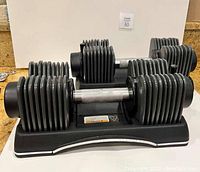 Two SA Gear adjustable dumbbells in black plastic base stands shown side by side on countertop.