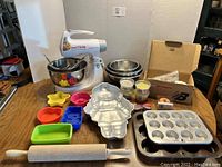 Overall view of Sunbeam mixer with multiple baking accessories on wooden table including bowls, cake molds, cupcake pans, rolling pin