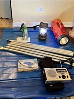 Overview of lot showing foldable camp table, multiple flashlights including large red flashlight, small green flashlight, lantern, folded metal rods, crank chargeable flashlight box, and L.L. Bean weather radio.
