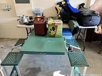 Wide shot of foldable kids picnic table with green seats and metal legs, assortment of cookware and camping equipment on table behind