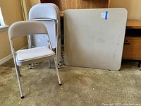 Photo showing 3 folded chairs and the card table standing upright, all light-colored and set on carpeted floor.