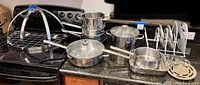 Photo showing the full set of stainless steel cookware items including pots, skillets, and racks arranged on kitchen counter and stove.