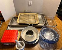 Various baking pans, cookie sheets, glass mixing bowls, baking stone with rack and handle, mortar and pestle spice grinder, and fabric pot holders displayed on a wooden table.