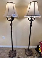 Two brown finish European style floor lamps side by side showing front view with beige shades and fringe trim.