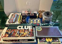 Photo of boxed games Clue (two editions), Monopoly, Scrabble with a wooden Scrabble board, multiple decks of playing cards, bingo markers, poker chip holder, and playing mat.