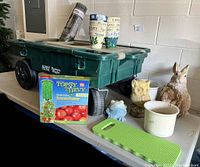Photo of green plastic garden cart with wheels, several pots including herb pots, and a Topsy Turvy tomato planter kit box on table