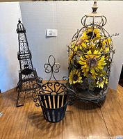 Three wrought iron decorative items on wood floor with white background: Eiffel Tower cork holder, bird cage floral display with sunflowers, and wall art/mail holder basket.