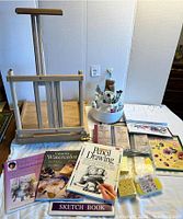 Wooden easel, paint brushes in caddy, oil paints tubes, instruction books, watercolor kits, and a spiral-bound sketchbook arranged on a table.