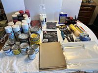 Full view of assorted paint cans, brushes, sandpaper, drop cloths, rollers, and tape laid out on table.