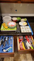 Top view of assorted plates, bowls, small dishes, paper napkins, and kitchen knives and utensils on the counter with two drawers open showing spoons, forks, knives and other kitchen utensils.