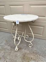 Front-side view of off white round painted accent table with scrollwork base outdoors on pavement.