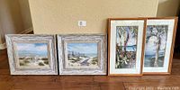 Four framed tropical art prints arranged on wooden floor: two with white washed wood frames showing beach scenes and two with bamboo style frames showing palm trees.