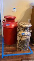 Two vintage metal milk containers side by side on a wooden floor against a beige wall. Red container is taller and in better condition, the white and black container is distressed.