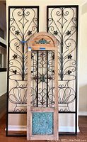 Three decorative panels including two black metal scroll panels and one wood and metal mixed panel with a curved top and blue painted metal floral design in the center.