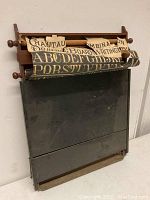 Front view of antique wooden school chalkboard with rolled alphabet lettering diagram partially unfurled above writing surface.