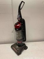 Full view of Hoover WindTunnel Rewind upright bagless vacuum showing vacuum base with wheels and handle, hose attached to side with brush accessory.