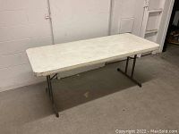 Side view showing full length of white plastic banquet table with metal legs, standing against a wall on a concrete floor.