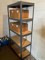 Full view of the metal shelving unit showing all four shelves with some boxes on upper shelves and empty lower shelves, positioned against a white concrete wall.