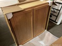 Photo of kitchen wall cabinet visible through torn open cardboard box, showing two wooden doors with raised panels.