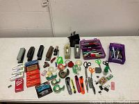 Overview of entire lot showing staplers, tape dispensers, utility knives, scissors, boxes of staples, pens in purple tray, and small tools on a white table.