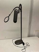 Full view of OttLite black desk lamp with flexible magnifier arm on white table against beige wall.