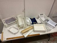 Wide shot showing all plastic baskets and organizers arranged on a table, mostly white with various sizes and shapes, including one with purple insert