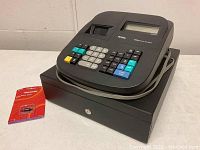 Front and side view of black Royal 500dx electronic cash register on closed cash drawer, with rolled cord and red ink roll cartridge nearby