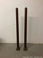 Front view of two tall antique wooden columns standing side by side against a plain wall, showing full height and square bases.