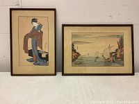 Photo showing two framed Japanese art pieces: a vertical ukiyo-e print of a woman holding a kimono, and a horizontal watercolor of a waterside village with boats.