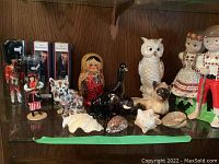 Photo showing Ukrainian boy and girl dolls, British Airways dolls in their original boxes, ceramic cat figurines, owl, black duck, shells, and a Russian nesting doll on a reflective surface against a wood background.